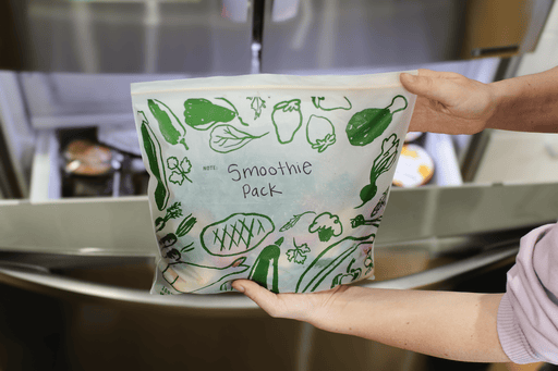 Person holding a smoothie pack inside a food storage bag with green vegetable illustrations in a kitchen setting
