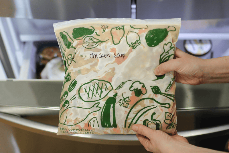 Hand holding a resealable food wrap package of 'Miso Chicken Soup' with plant illustrations near a freezer