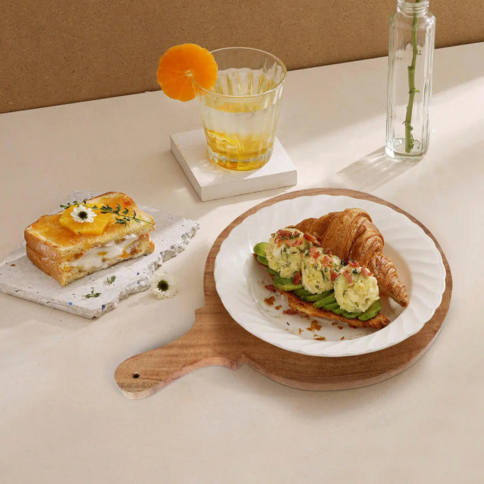 Breakfast setting with croissant, avocado toast, and glass of orange juice on a wooden board on a white table.